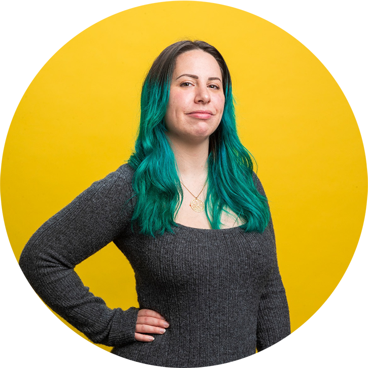 Portrait photo of Betsy smiling with her hand on her hip and a yellow background
