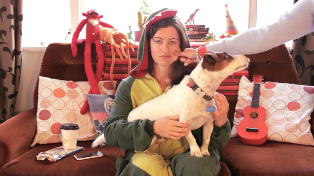 A person sitting on a couch, wearing a onesie with a dead pan expression while holding a dog on her lap.
