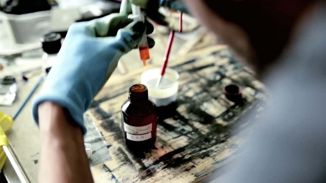 A closeup photo of an artist wearing gloves, mixing colours with a syringe and glass bottle