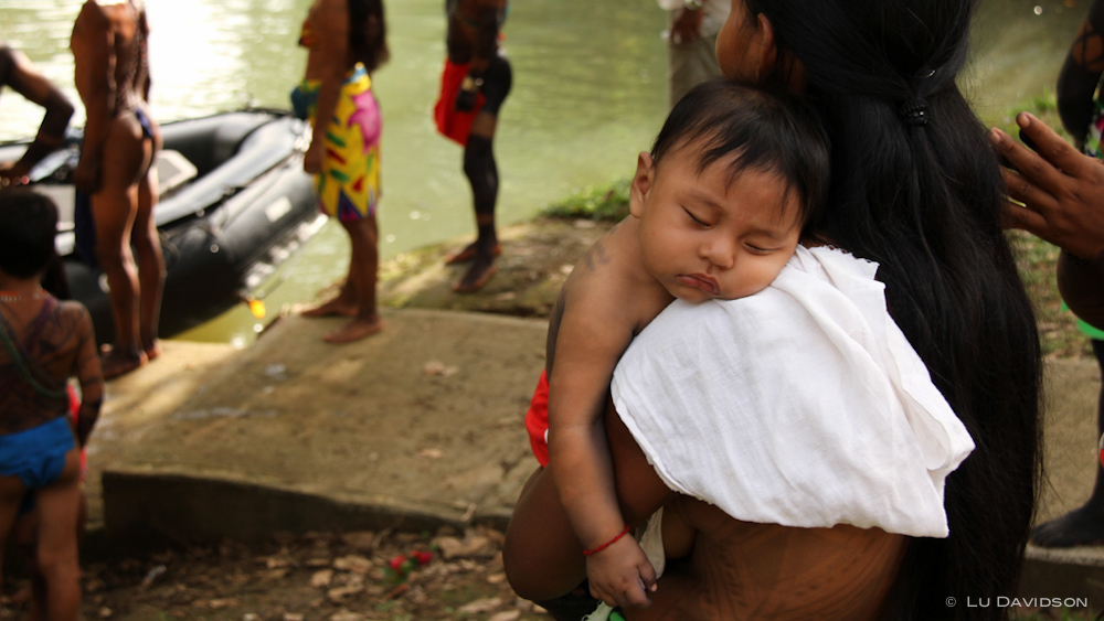 A small child sleeping while being held by the mother, with people standing by a river in the background.